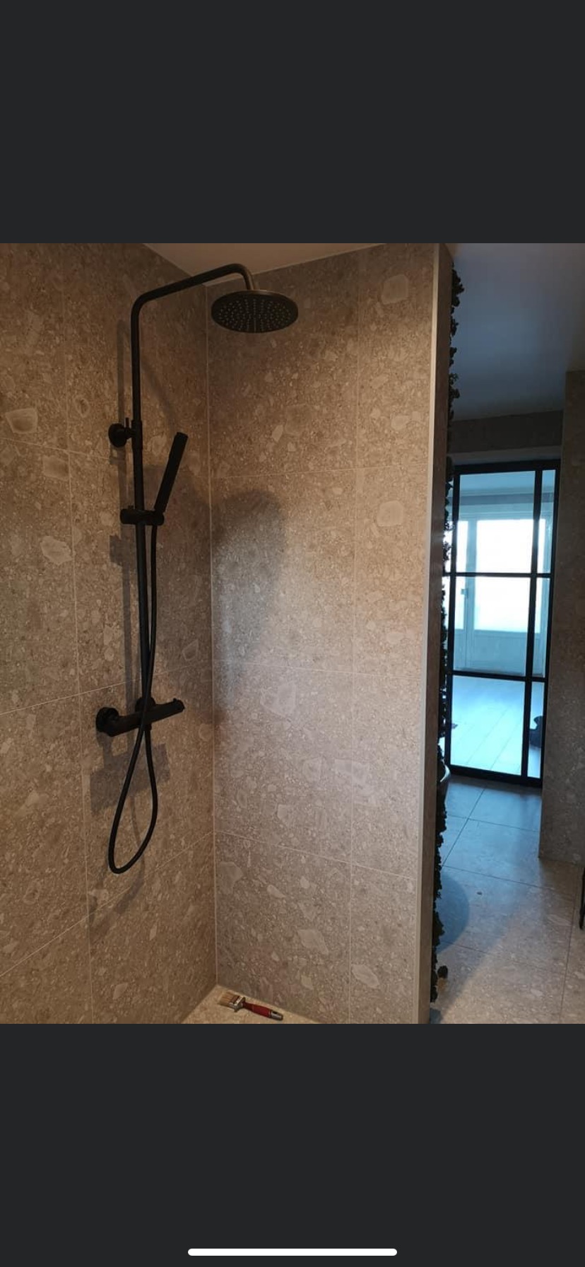 Nowoczesna kabina prysznicowa z czarną armaturą na tle beżowych płytek imitujących kamień, widoczny pędzel na podłodze i fragment czarnej ramy drzwi w tle.