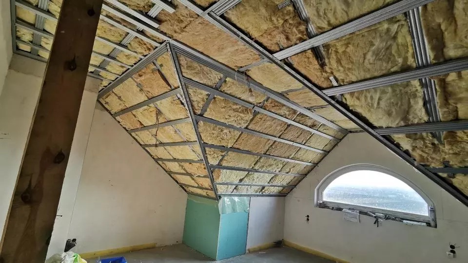 Poddasze w trakcie izolacji: wełna mineralna w metalowym stelażu, widoczna drewniana belka, okrągłe okno i fragment płyty gipsowo-kartonowej.