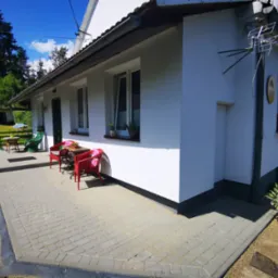 Wyłożony szarą kostką brukową taras przed białym budynkiem z anteną satelitarną, czerwone krzesła i stolik na tarasie, doniczki z kwiatami w oknach i przy wejściu.