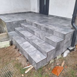 TomBud - Wykończone schody wejściowe do domu, obłożone szarą, kwadratową glazurą imitującą kamień, z widocznym fragmentem elewacji i rynny spustowej, teren wokół schodów częściowo zagospodarowany,...