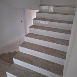 TomBud - Wykończone schody wewnętrzne z szarych płytek imitujących marmur, z białymi podstopnicami, widok z góry na dół, w tle surowa ściana.