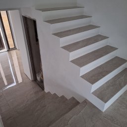 TomBud - Wykończone schody wewnętrzne z płytkami imitującymi marmur, z widoczną podłogą z tego samego materiału, oraz przygotowane wejście do pomieszczenia w stanie surowym.