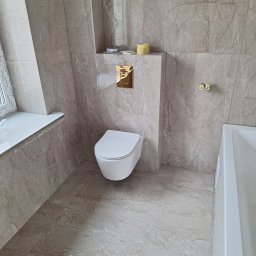 TomBud - Nowoczesna łazienka z beżowymi płytkami imitującymi marmur na ścianach i podłodze, wisząca toaleta z złotym przyciskiem spłukującym, biała wanna i okno z białym parapetem.