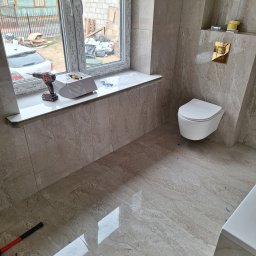 TomBud - Wisząca toaleta w wykończonej łazience z beżowymi płytkami imitującymi marmur, widoczny akumulatorowy wkrętak na parapecie, młotek i miarka na podłodze, okno z widokiem na budynek w trakcie budowy.