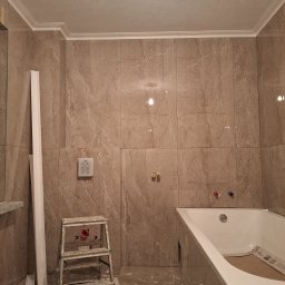 TomBud - Łazienka w trakcie wykańczania: marmurowe płytki, biała wanna, drabina malarska, listwy przypodłogowe i sufitowe, przygotowane przyłącza wodne.