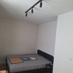 TomBud - Pokój po odświeżeniu, z pomalowanymi ścianami w odcieniach bieli i szarości, oświetleniem szynowym z czarnymi reflektorami na suficie, podłogą z parkietu ułożonego w jodełkę i łóżkiem z trzema...