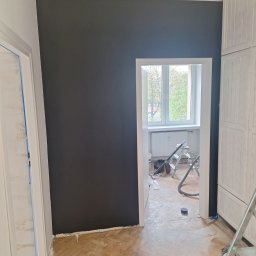 TomBud - Pomieszczenie w trakcie odświeżania: ściana pomalowana na ciemny grafit, fragment podłogi z parkietu, widoczne narzędzia malarskie, drabina malarska i odkurzacz w tle.