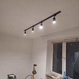 TomBud - Pokój w trakcie odświeżania: świeżo pomalowane, białe ściany, czarna szyna z reflektorami sufitowymi, okno zabezpieczone taśmą malarską, na podłodze wiadro z farbą i lampa warsztatowa.