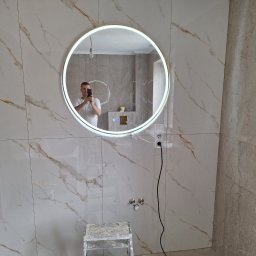 TomBud - Łazienka po remoncie z dużymi płytkami imitującymi marmur na ścianach, okrągłe lustro z podświetleniem LED, widoczne narzędzia i drabina malarska, odbicie fotografa w lustrze.