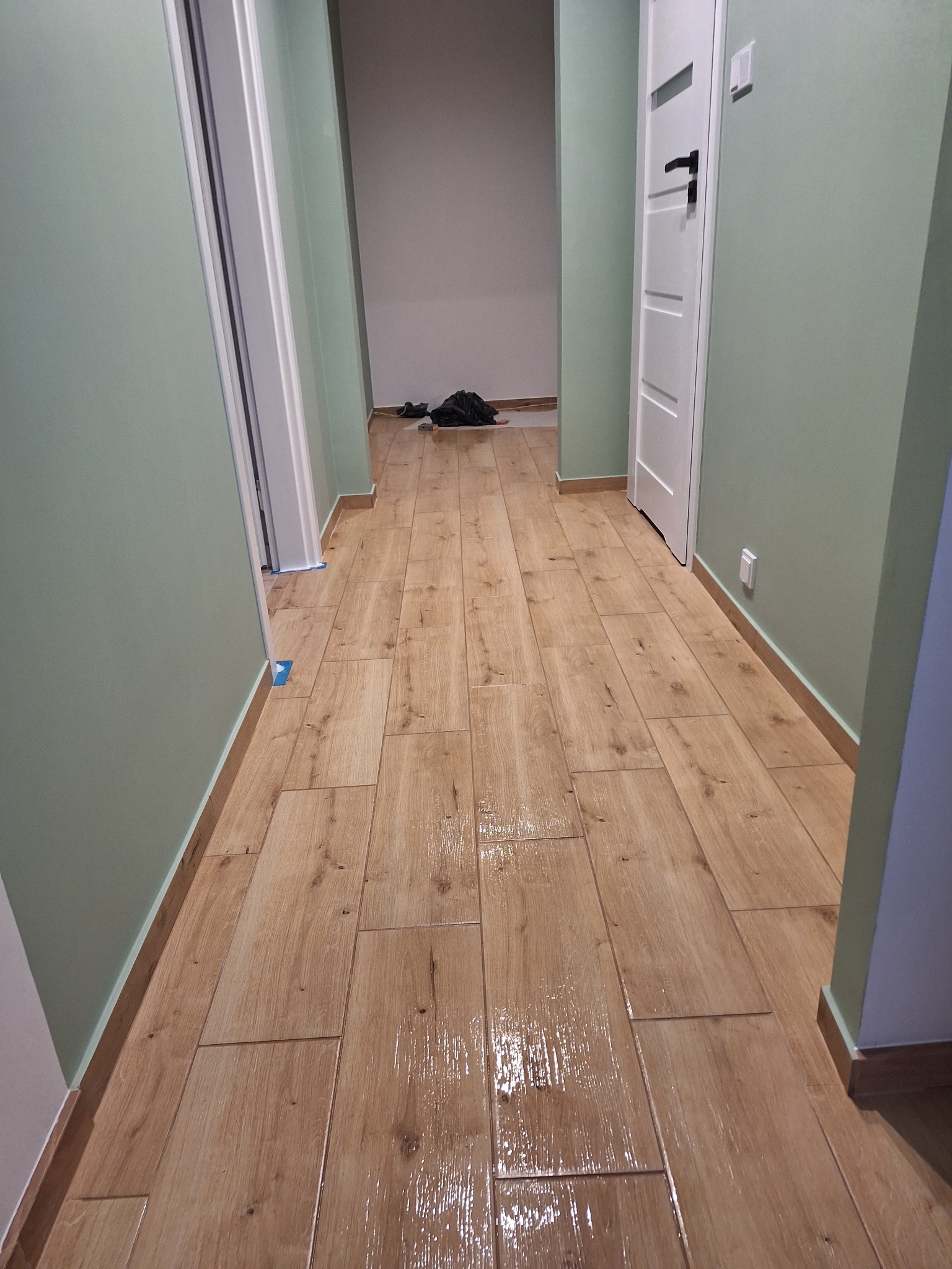 Korytarz po ułożeniu podłogi z płytek imitujących drewno, świeżo umyte, z widocznymi refleksami światła na powierzchni, ściany pomalowane na pistacjowy kolor.