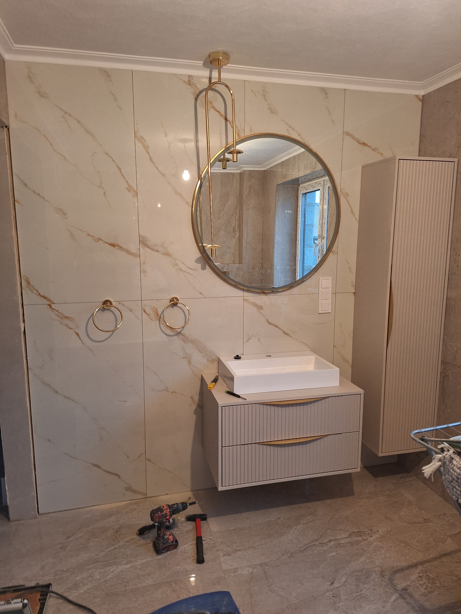 Nowoczesna łazienka w trakcie wykańczania: beżowe płytki imitujące marmur, okrągłe lustro w złotej ramie, wisząca szafka z umywalką, złote uchwyty na ręczniki, narzędzia na podłodze.