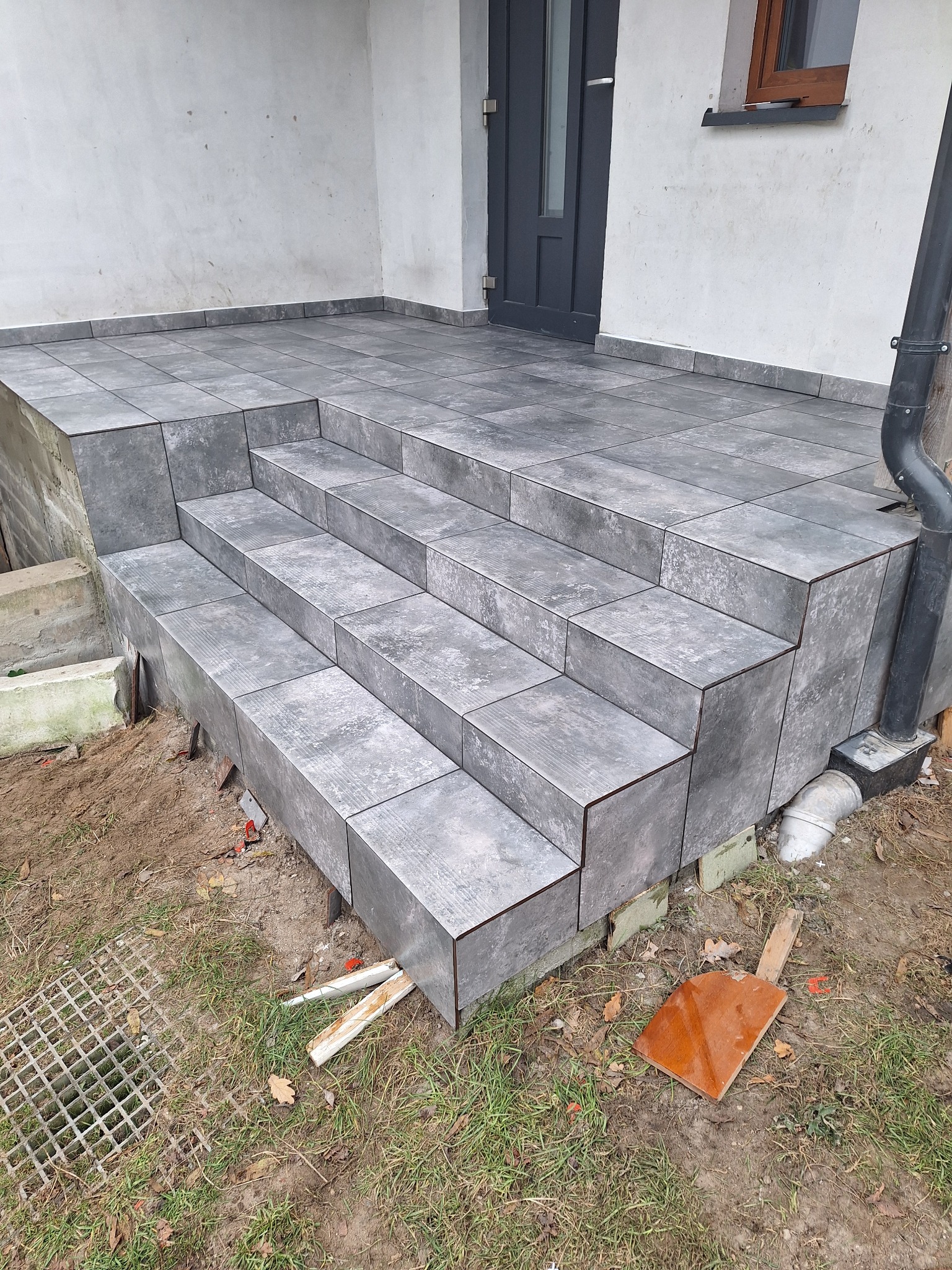 Wykończone schody wejściowe do domu, obłożone szarą, kwadratową glazurą imitującą kamień, z widocznym fragmentem elewacji i rynny spustowej, teren wokół schodów częściowo zagospodarowany,...