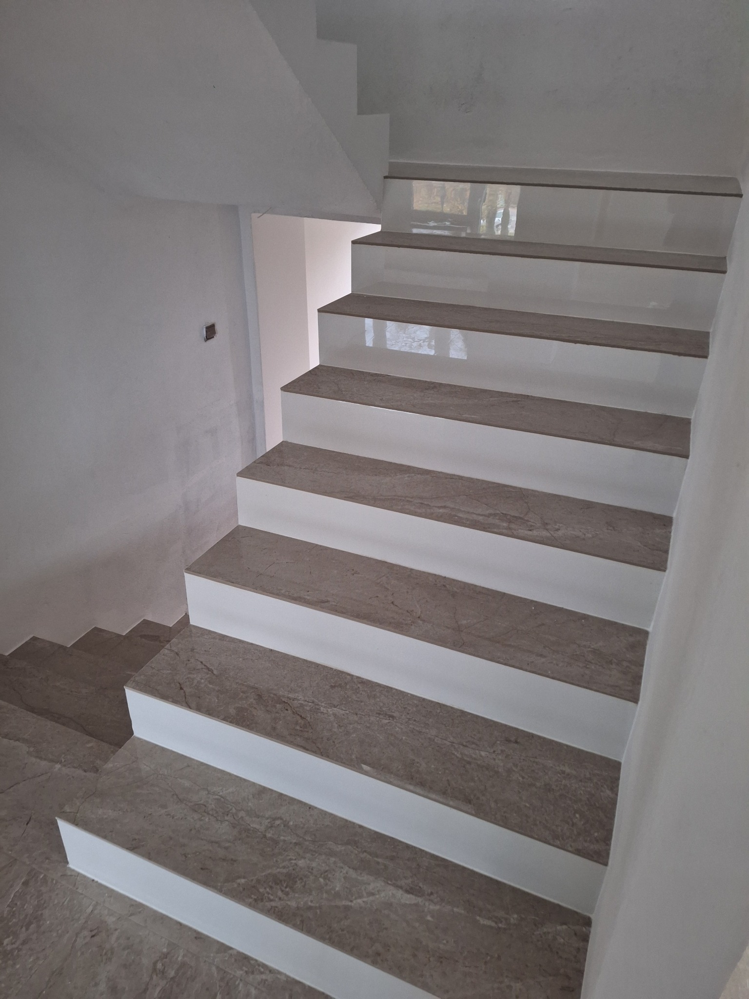 Wykończone schody wewnętrzne z szarych płytek imitujących marmur, z białymi podstopnicami, widok z góry na dół, w tle surowa ściana.