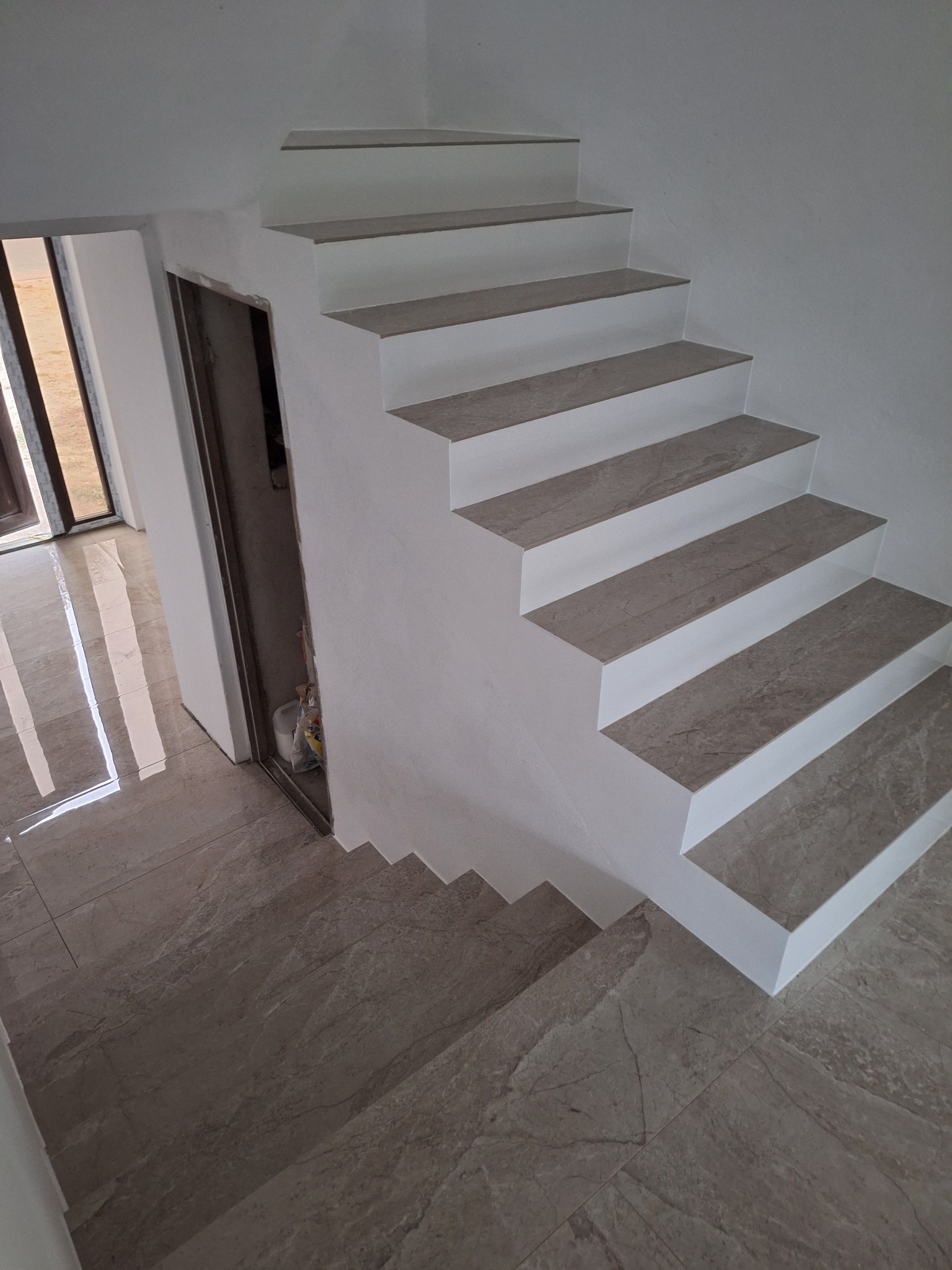 Wykończone schody wewnętrzne z płytkami imitującymi marmur, z widoczną podłogą z tego samego materiału, oraz przygotowane wejście do pomieszczenia w stanie surowym.