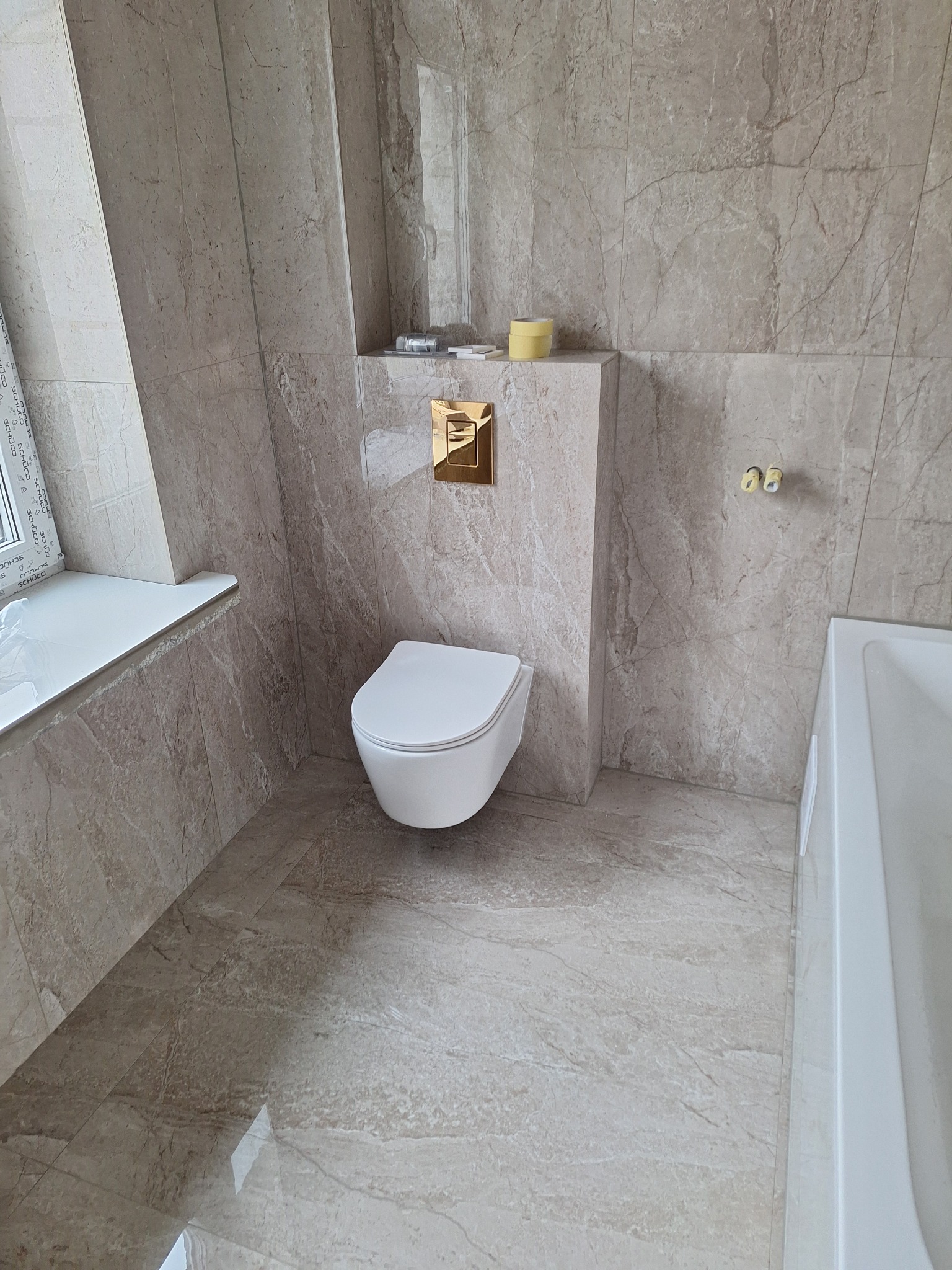 Nowoczesna łazienka z beżowymi płytkami imitującymi marmur na ścianach i podłodze, wisząca toaleta z złotym przyciskiem spłukującym, biała wanna i okno z białym parapetem.