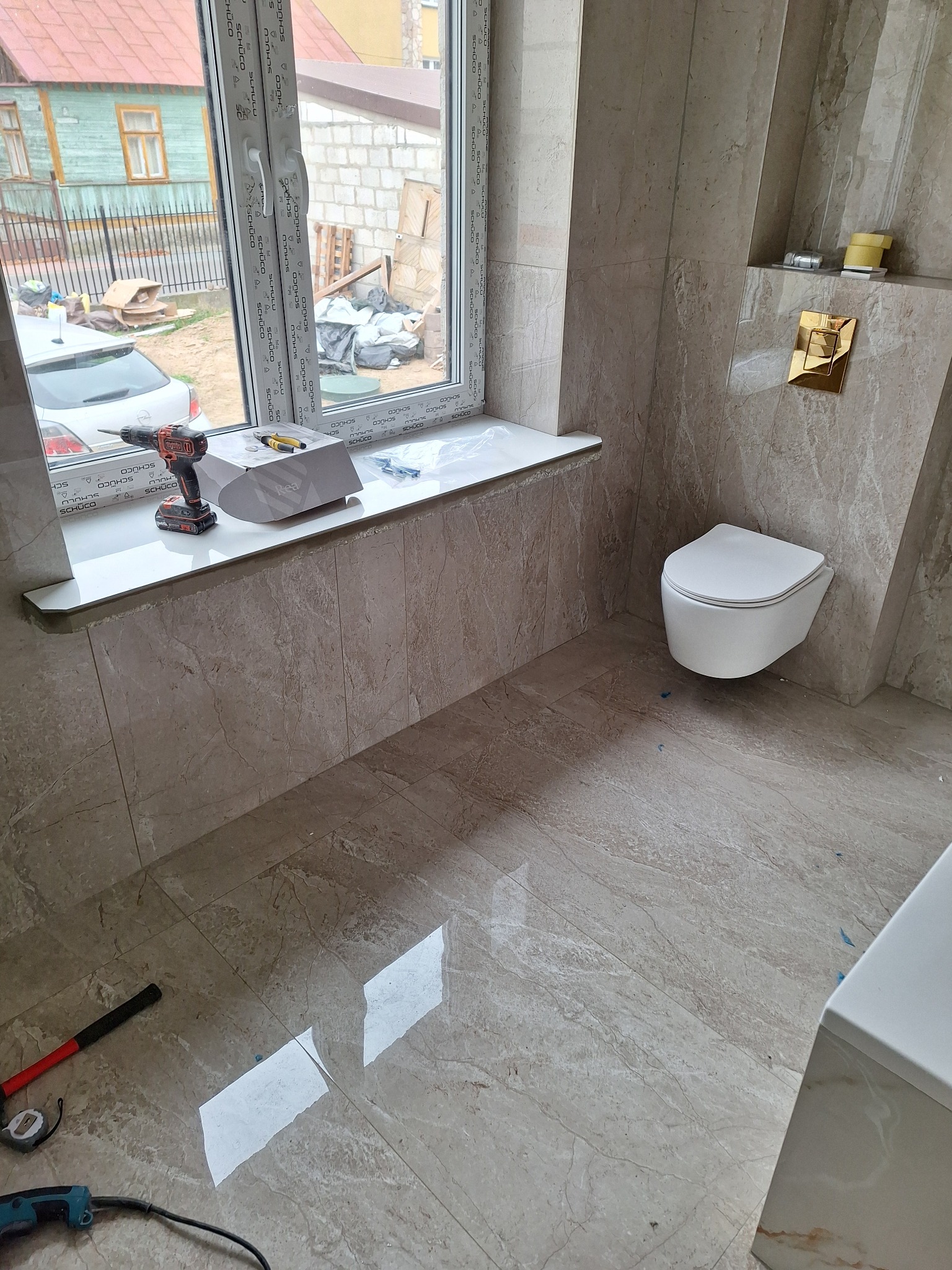 Wisząca toaleta w wykończonej łazience z beżowymi płytkami imitującymi marmur, widoczny akumulatorowy wkrętak na parapecie, młotek i miarka na podłodze, okno z widokiem na budynek w trakcie budowy.