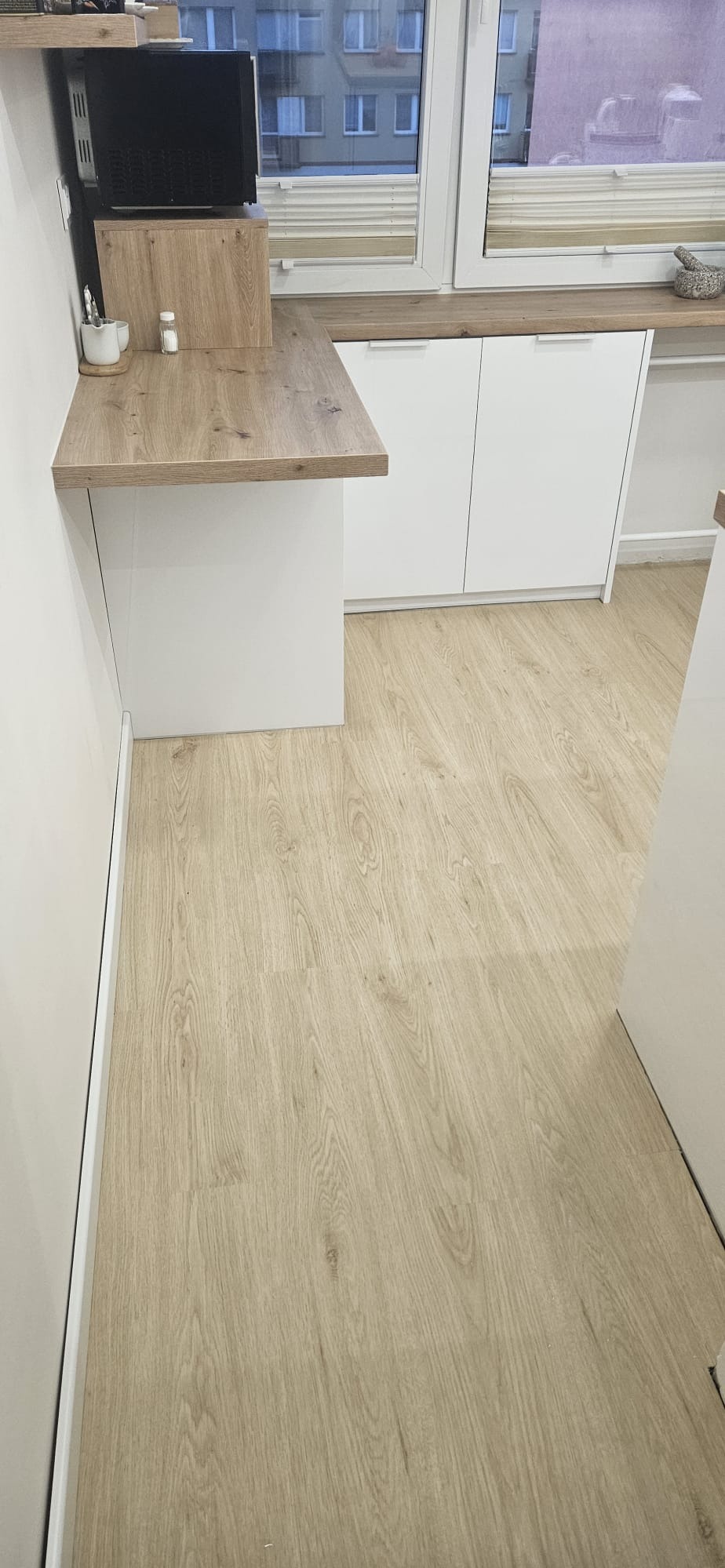 Jasne panele podłogowe z widoczną strukturą drewna, ułożone w nowej kuchni z białymi szafkami i blatem w odcieniu dębu.