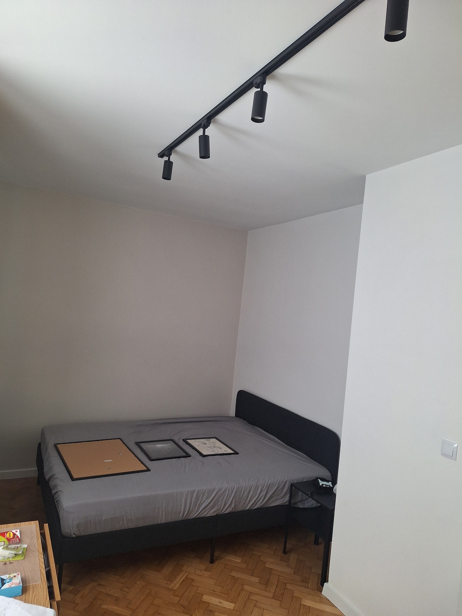 Pokój po odświeżeniu, z pomalowanymi ścianami w odcieniach bieli i szarości, oświetleniem szynowym z czarnymi reflektorami na suficie, podłogą z parkietu ułożonego w jodełkę i łóżkiem z trzema...