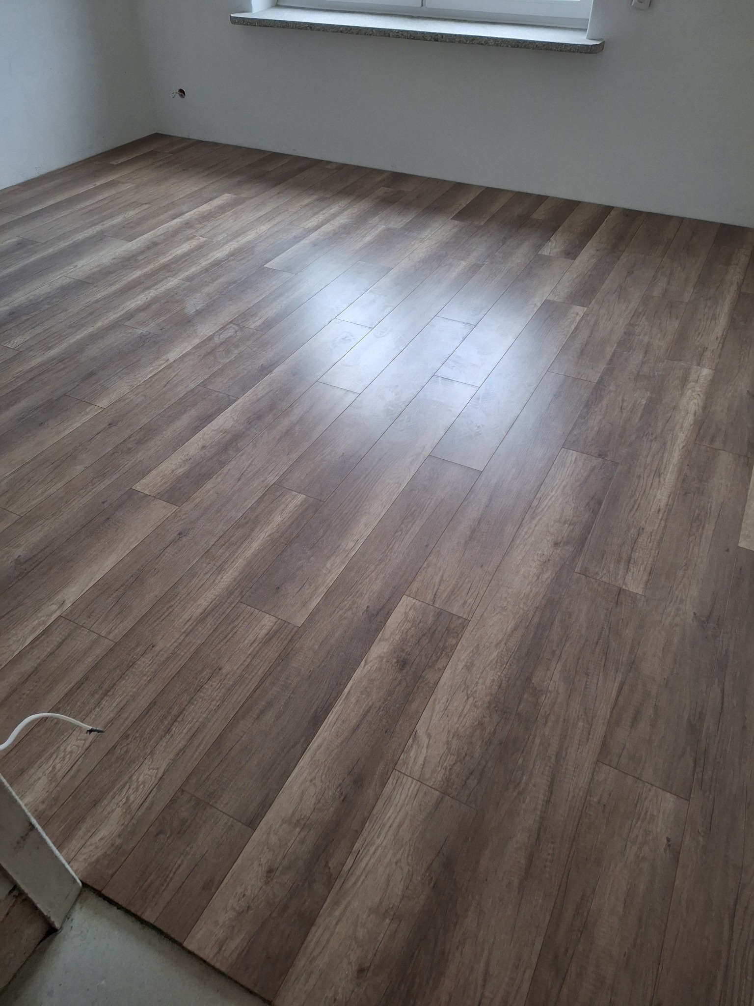 Nowo ułożone panele podłogowe w odcieniach brązu z widocznym fragmentem ściany i parapetem okiennym, widać odbicie światła na panelach.