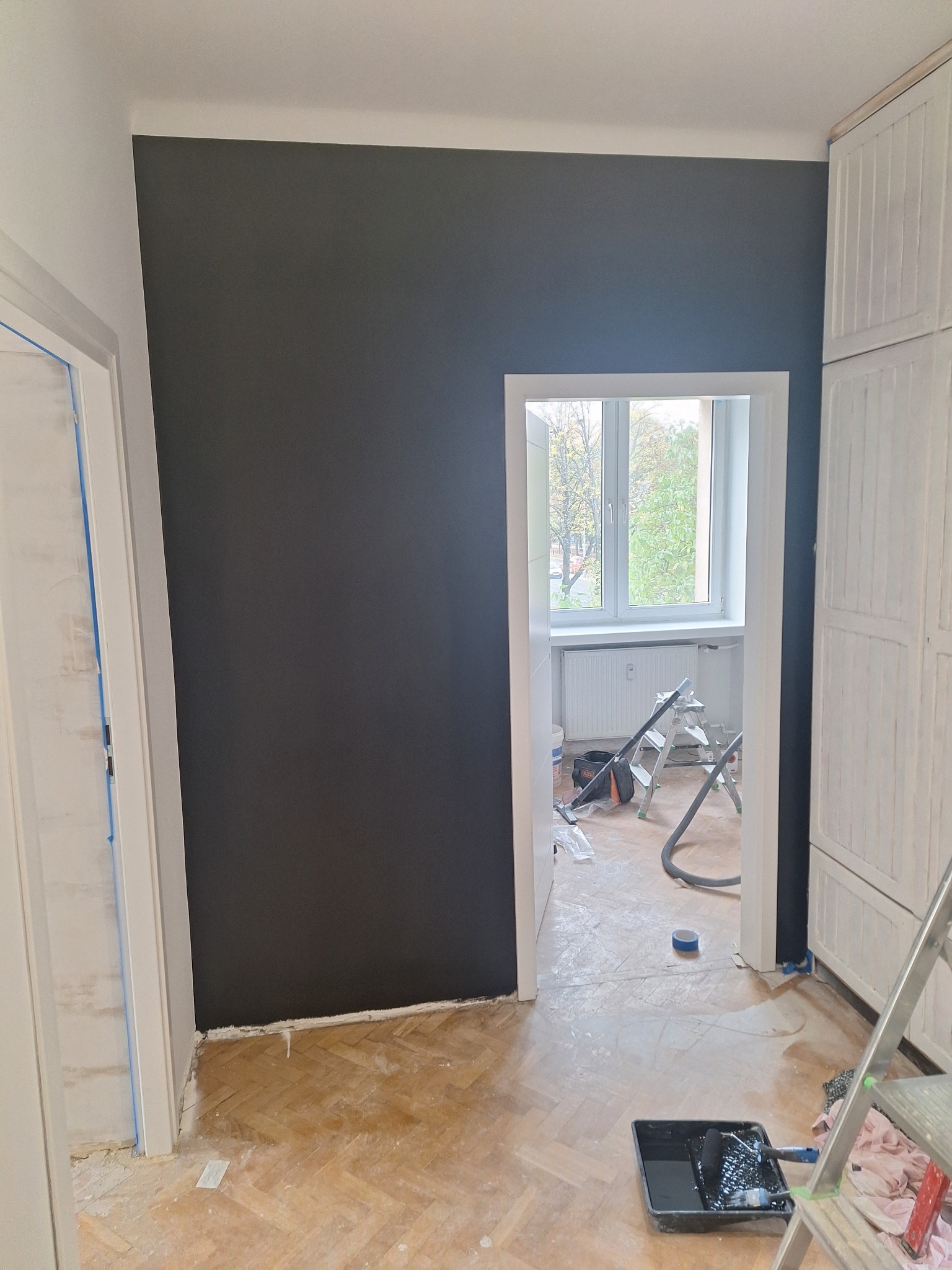 Pomieszczenie w trakcie odświeżania: ściana pomalowana na ciemny grafit, fragment podłogi z parkietu, widoczne narzędzia malarskie, drabina malarska i odkurzacz w tle.