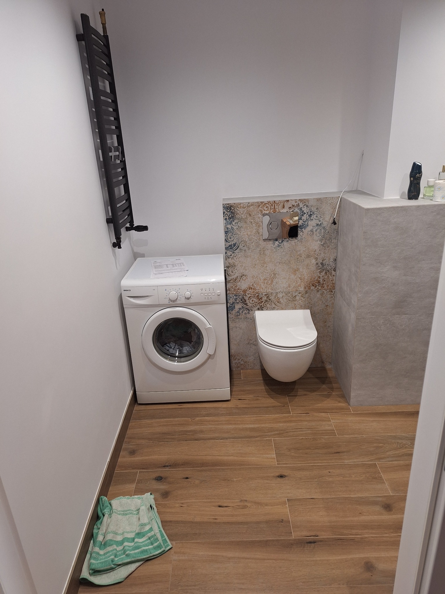 Nowoczesna łazienka z drewnopodobnymi płytkami podłogowymi, wiszącą toaletą, pralką i grzejnikiem drabinkowym. Dekoracyjna ściana nad toaletą z wzorzystymi płytkami. Na podłodze leży zielony ręcznik.