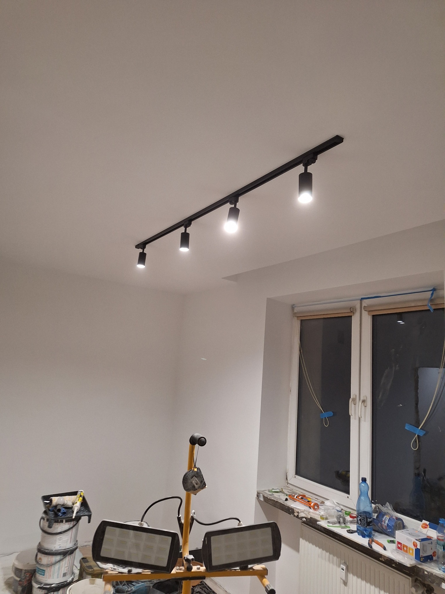Pokój w trakcie odświeżania: świeżo pomalowane, białe ściany, czarna szyna z reflektorami sufitowymi, okno zabezpieczone taśmą malarską, na podłodze wiadro z farbą i lampa warsztatowa.