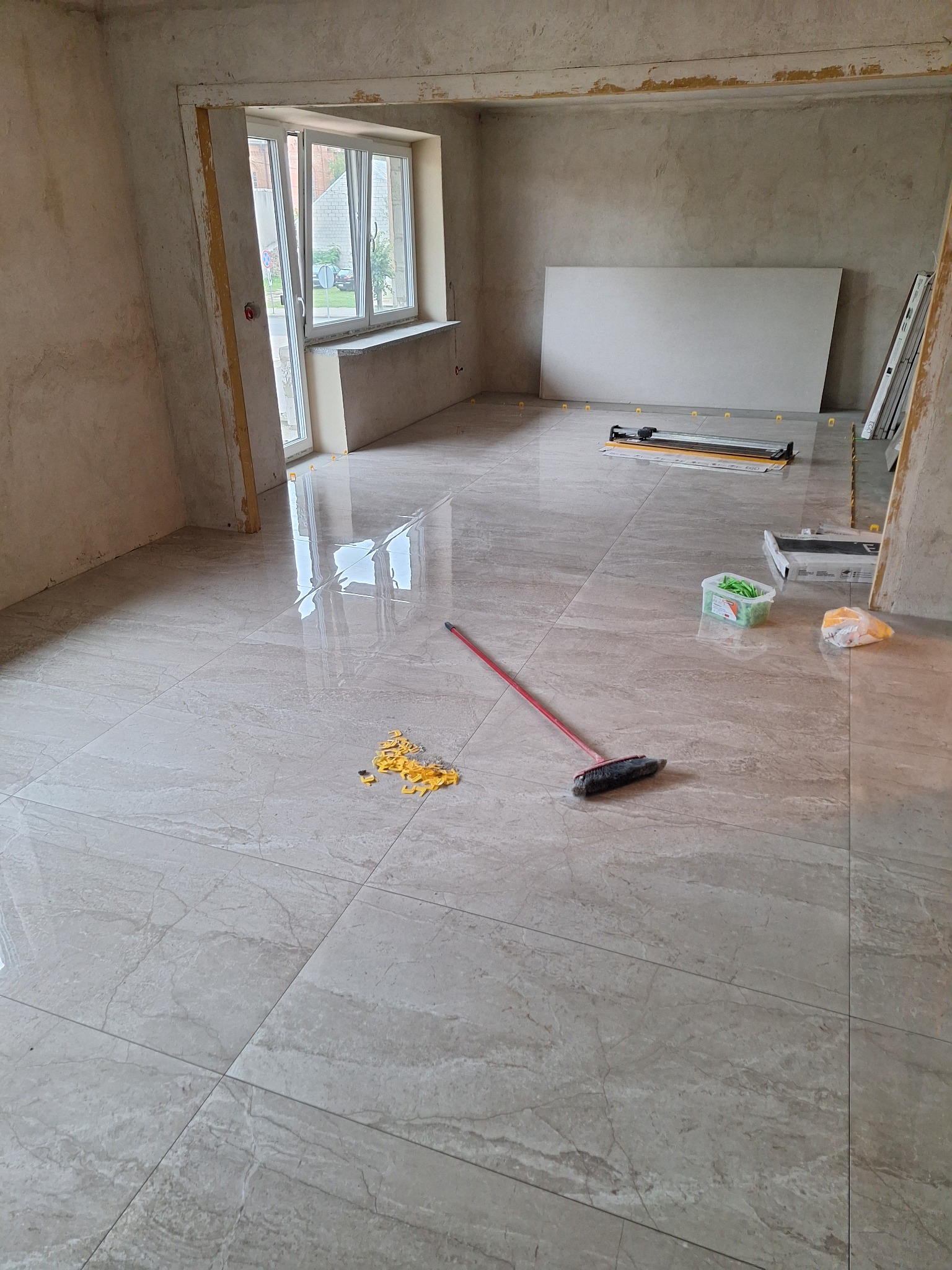 Błyszczące, beżowe płytki podłogowe imitujące marmur, ułożone w trakcie remontu, widoczne narzędzia glazurnicze, poziomica, zapas płytek, kliny dystansowe i szczotka do zamiatania.