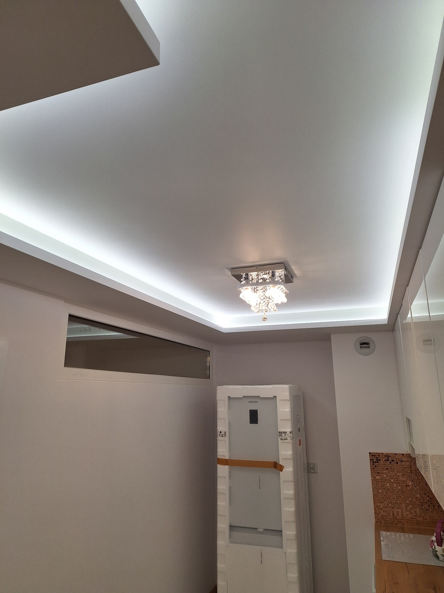 Biały sufit z podwieszanym oświetleniem LED i kryształowym żyrandolem w łazience, widoczna wentylacja, okno i lodówka w folii ochronnej.