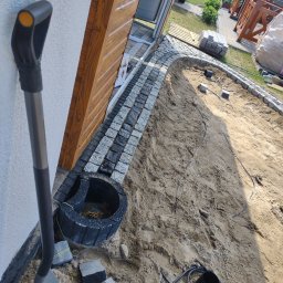 ER-BRUK - Przygotowanie podłoża pod ścieżkę z kostki granitowej, widoczna łopata, obrzeża z kostki i piasek.