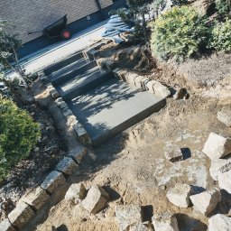 ER-BRUK - Wykonywanie schodów zewnętrznych z betonowych stopni obłożonych kostką granitową, widoczna taczka i węże na górze skarpy.