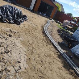 ER-BRUK - Wstępny etap tworzenia ścieżki z kostki granitowej na piaszczystym podłożu, widoczne narzędzia i materiały budowlane, w tle budynki gospodarcze.