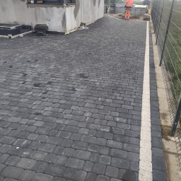 ER-BRUK - Ułożona, ciemnoszara kostka brukowa tworząca podjazd, z białym pasem oddzielającym od ogrodzenia z siatki. W tle widoczny budynek w trakcie wykończenia i pracownik w pomarańczowym ubraniu.
