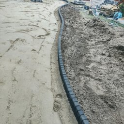 ER-BRUK - Przygotowanie podłoża pod ścieżkę z ciemnej kostki brzegowej; widoczne warstwy piasku i ziemi, oraz pracownicy w tle