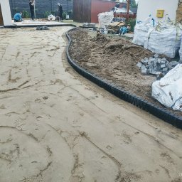 ER-BRUK - Przygotowanie podłoża pod kostkę brukową z ułożonym obrzeżem z tworzywa, widoczne worki z materiałem i ekipa budowlana w tle.