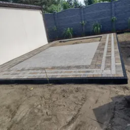 Fragment podwórka z wykończoną obwódką z kostki brukowej i wysypanym drobnym kamieniem, widoczne zapasy kostki i świeżo posadzone rośliny wzdłuż ogrodzenia.