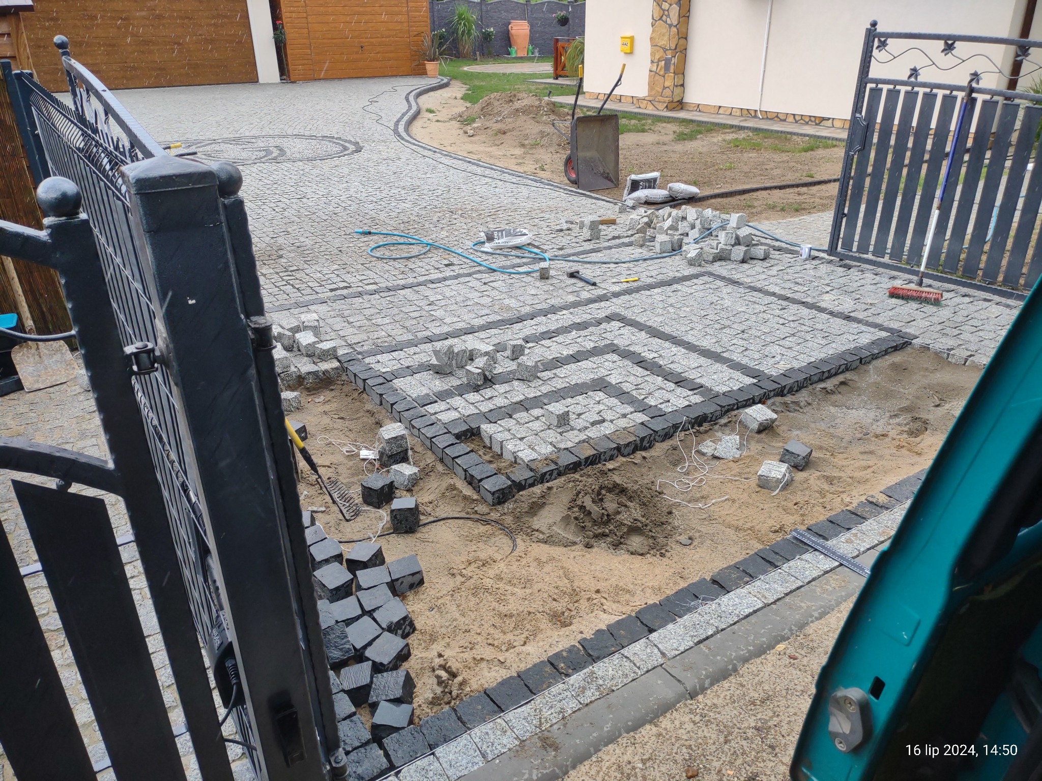 Układanie wzoru z jasnoszarej i czarnej kostki granitowej na podjeździe, widoczne narzędzia i materiały budowlane, brama w tle.