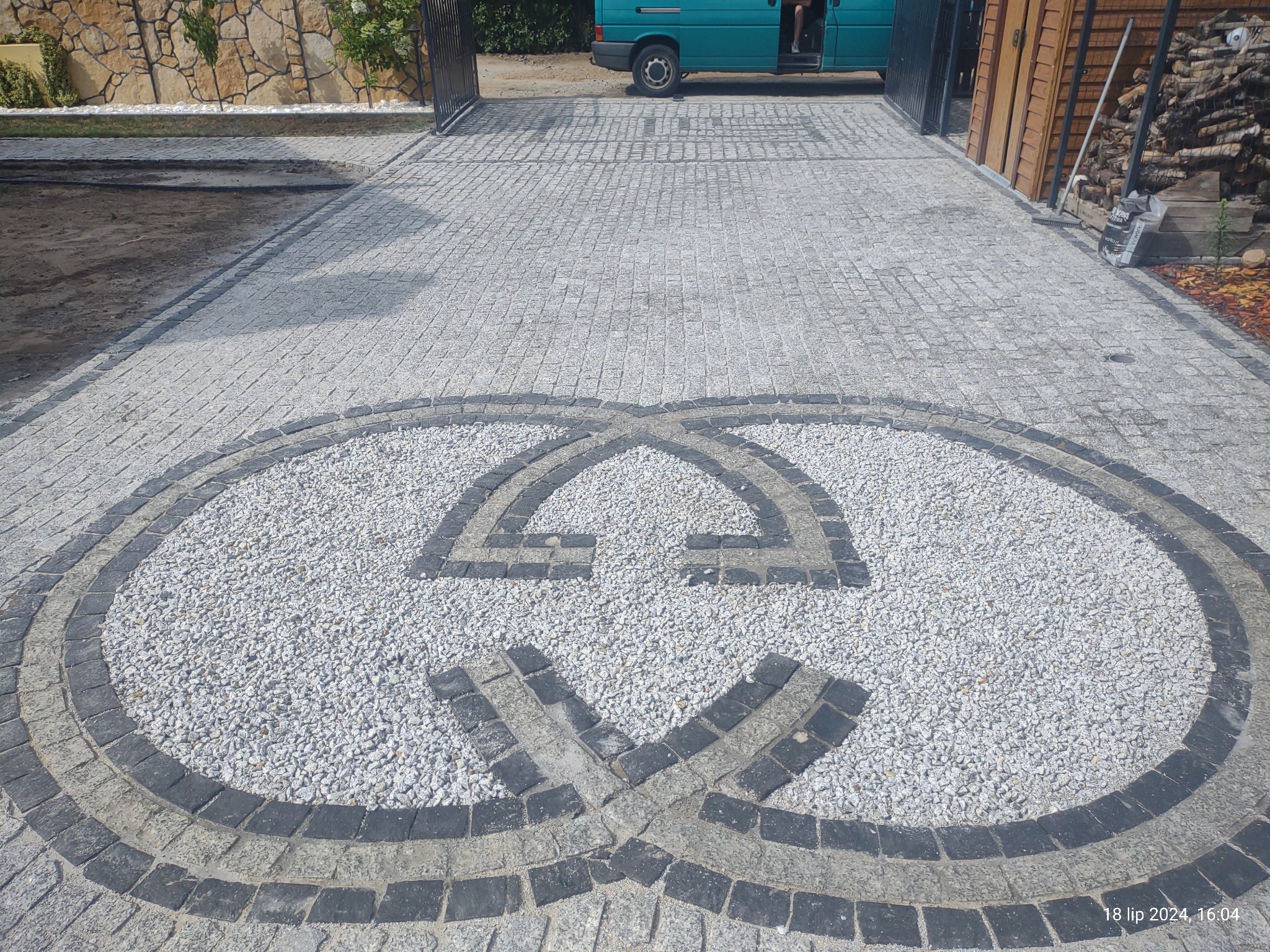 Szeroki podjazd z szarej kostki granitowej z dekoracyjnym wzorem z ciemnej kostki i białego grysu, na tle zielonego busa.