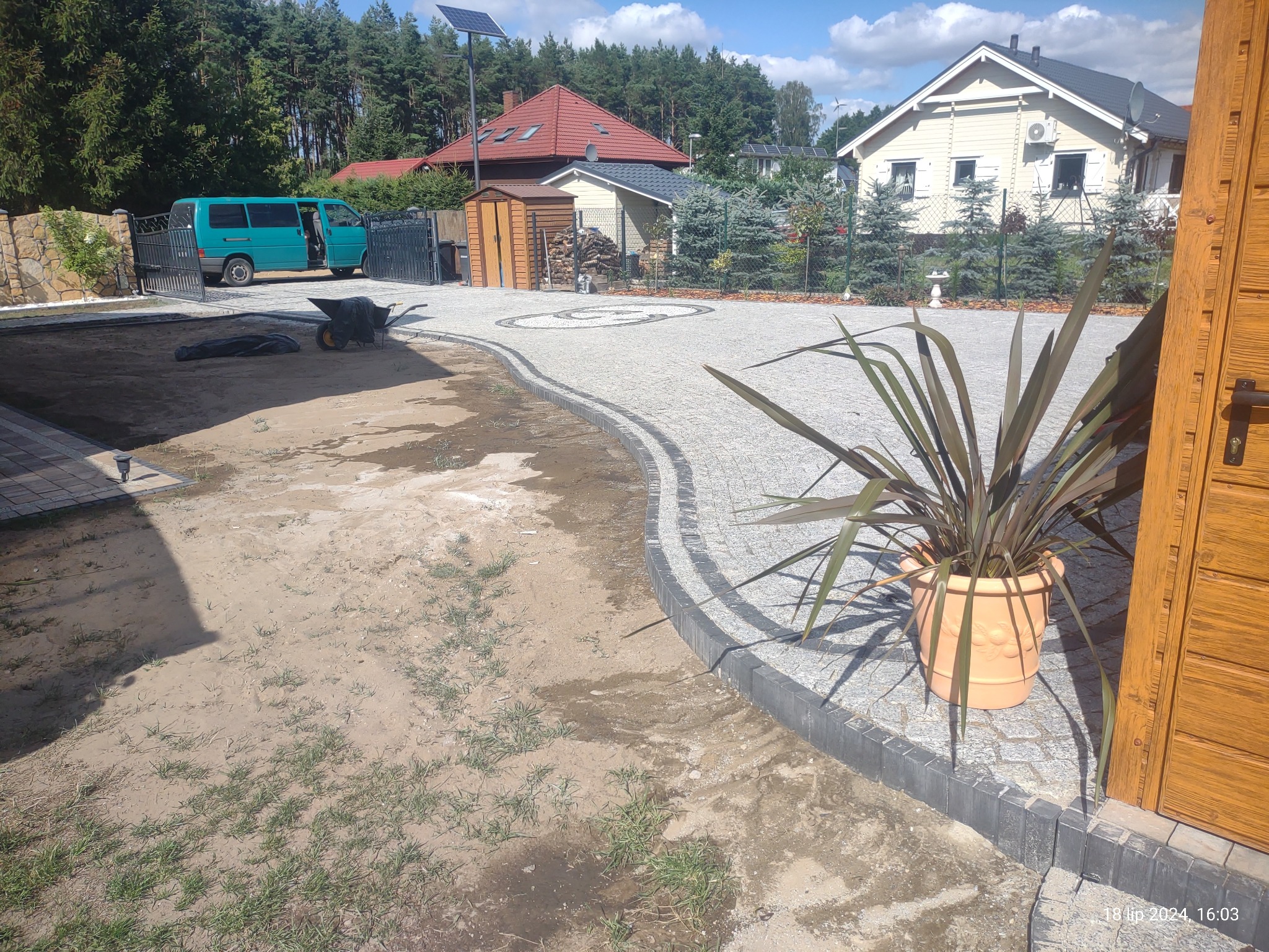 Ułożona ścieżka z jasnoszarej kostki granitowej z ciemnoszarym obrzeżem, widok z poziomu gruntu na ogród z domami w tle, taczka i bus na posesji.