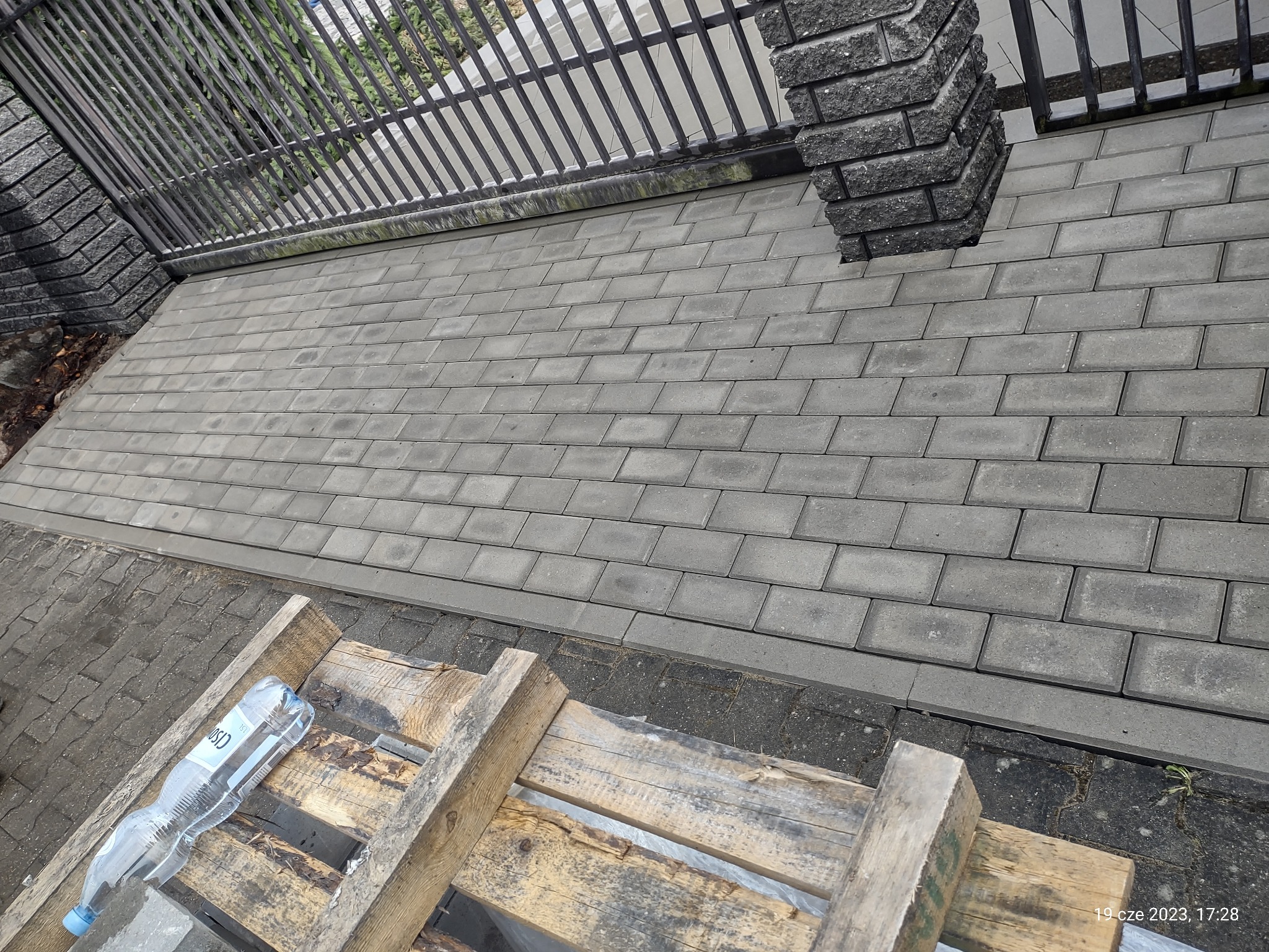 Świeżo ułożona szara kostka brukowa przy metalowym ogrodzeniu, widoczna butelka wody na drewnianym rusztowaniu, perspektywa z góry.