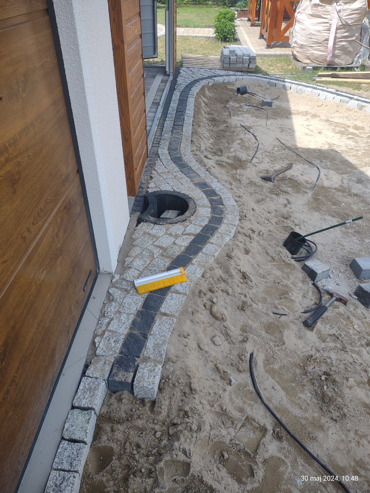 Układanie falującej ścieżki z kostki granitowej w piasku, widoczny żółty zmiotak, narzędzia i worki z materiałem w tle.