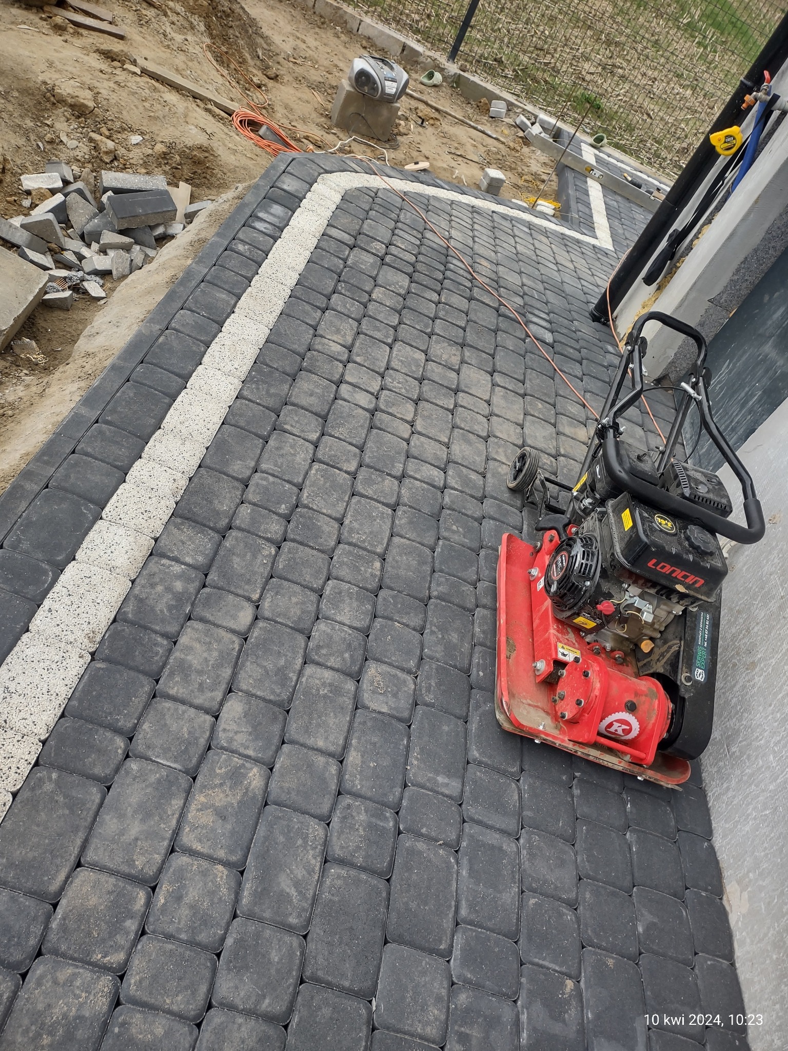 Częściowo ułożona, ciemnoszara kostka brukowa z białym obramowaniem, zagęszczarka wibracyjna stoi na ułożonej części, widoczne zapasy kostki i piasku w tle.