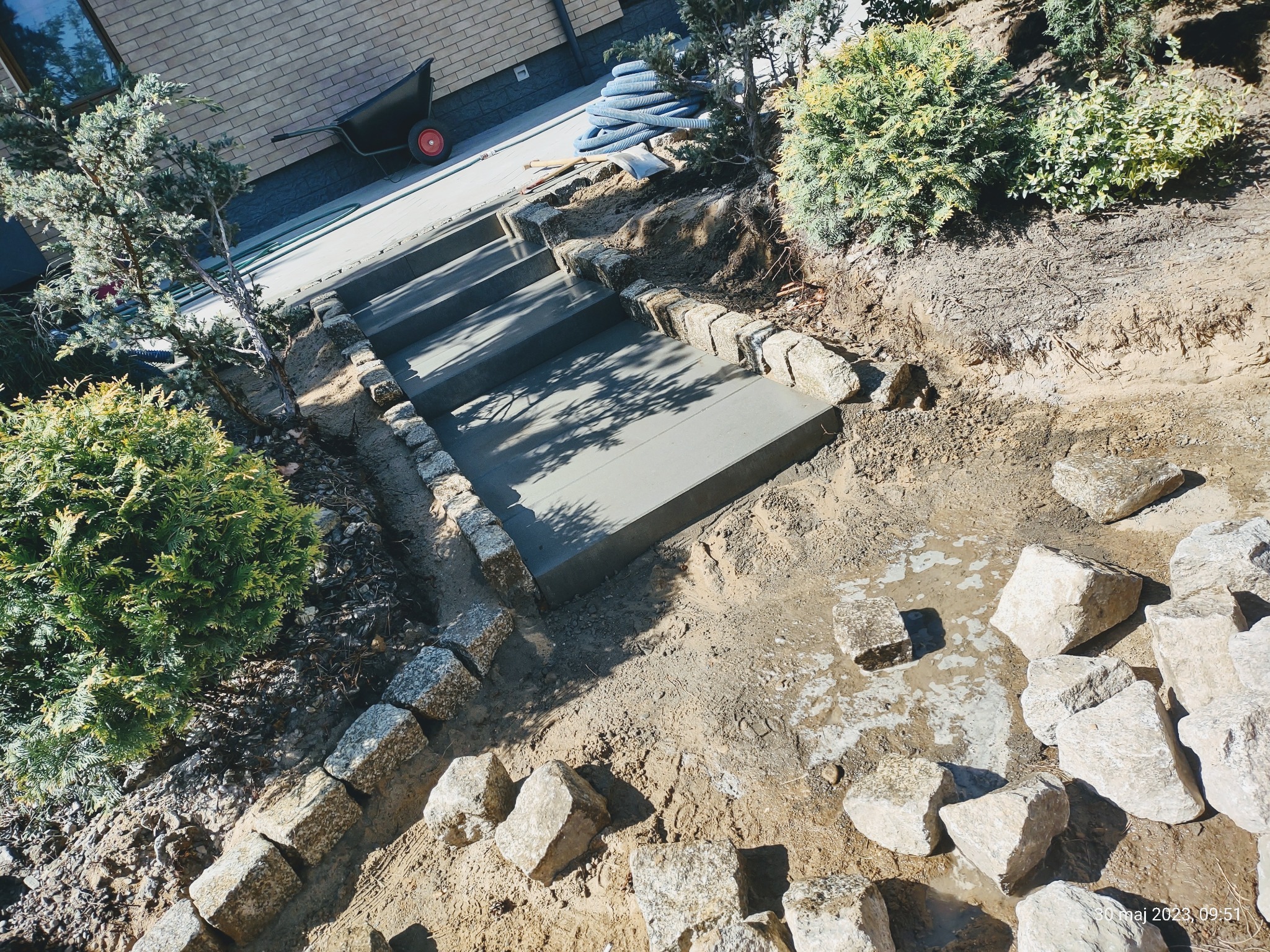 Wykonywanie schodów zewnętrznych z betonowych stopni obłożonych kostką granitową, widoczna taczka i węże na górze skarpy.