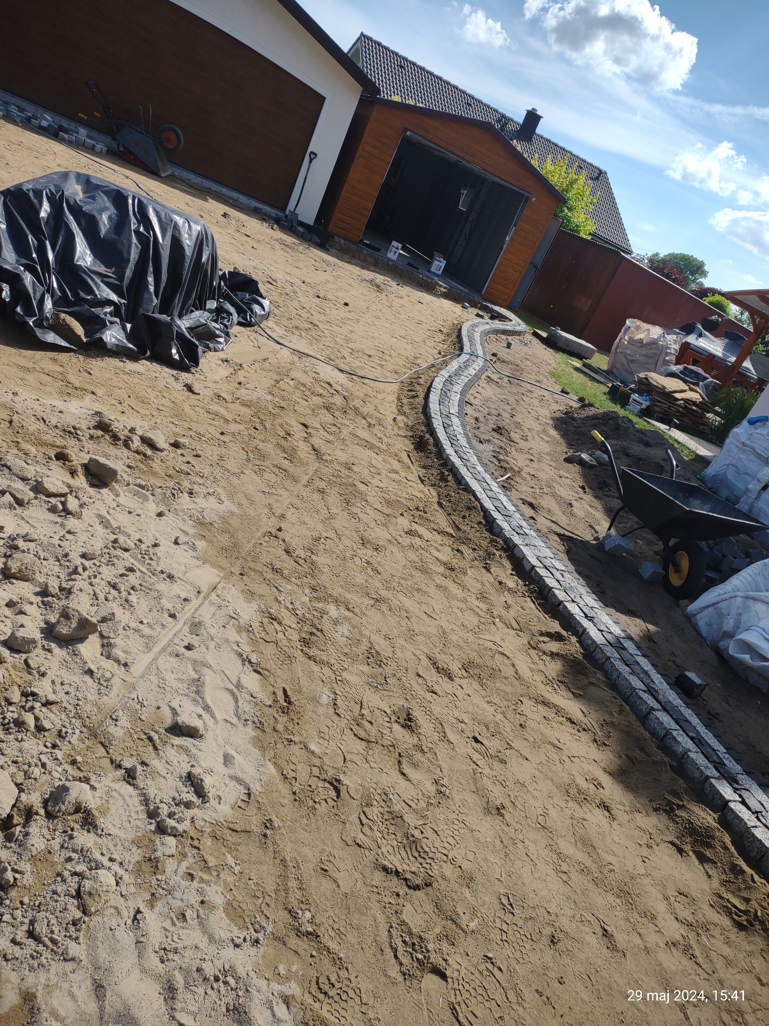 Wstępny etap tworzenia ścieżki z kostki granitowej na piaszczystym podłożu, widoczne narzędzia i materiały budowlane, w tle budynki gospodarcze.