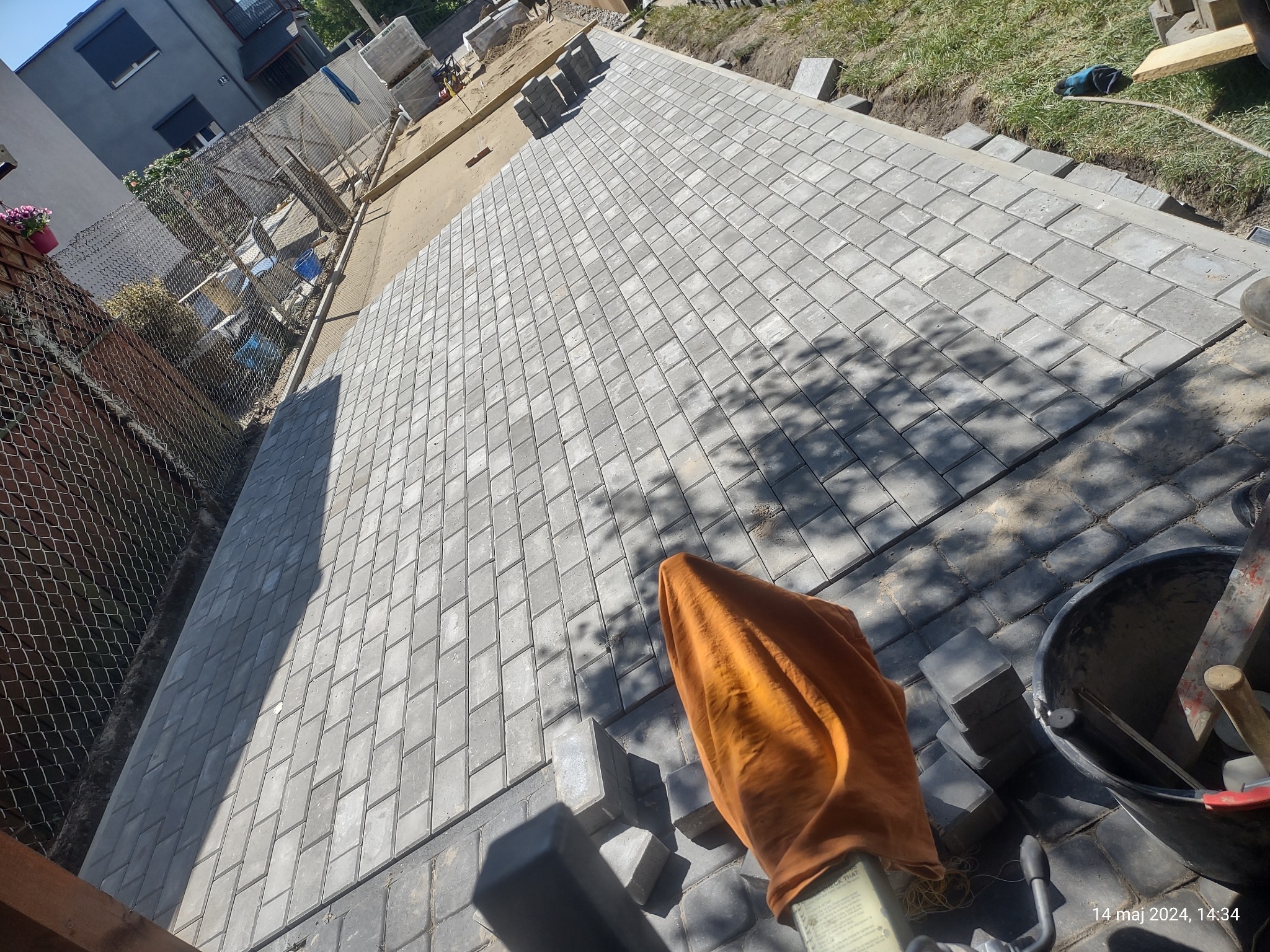 Ułożona szara kostka brukowa w romby, widok z góry, narzędzia w wiadrze i pomarańczowa szmatka w prawym dolnym rogu, w tle ogrodzenie i budynek.