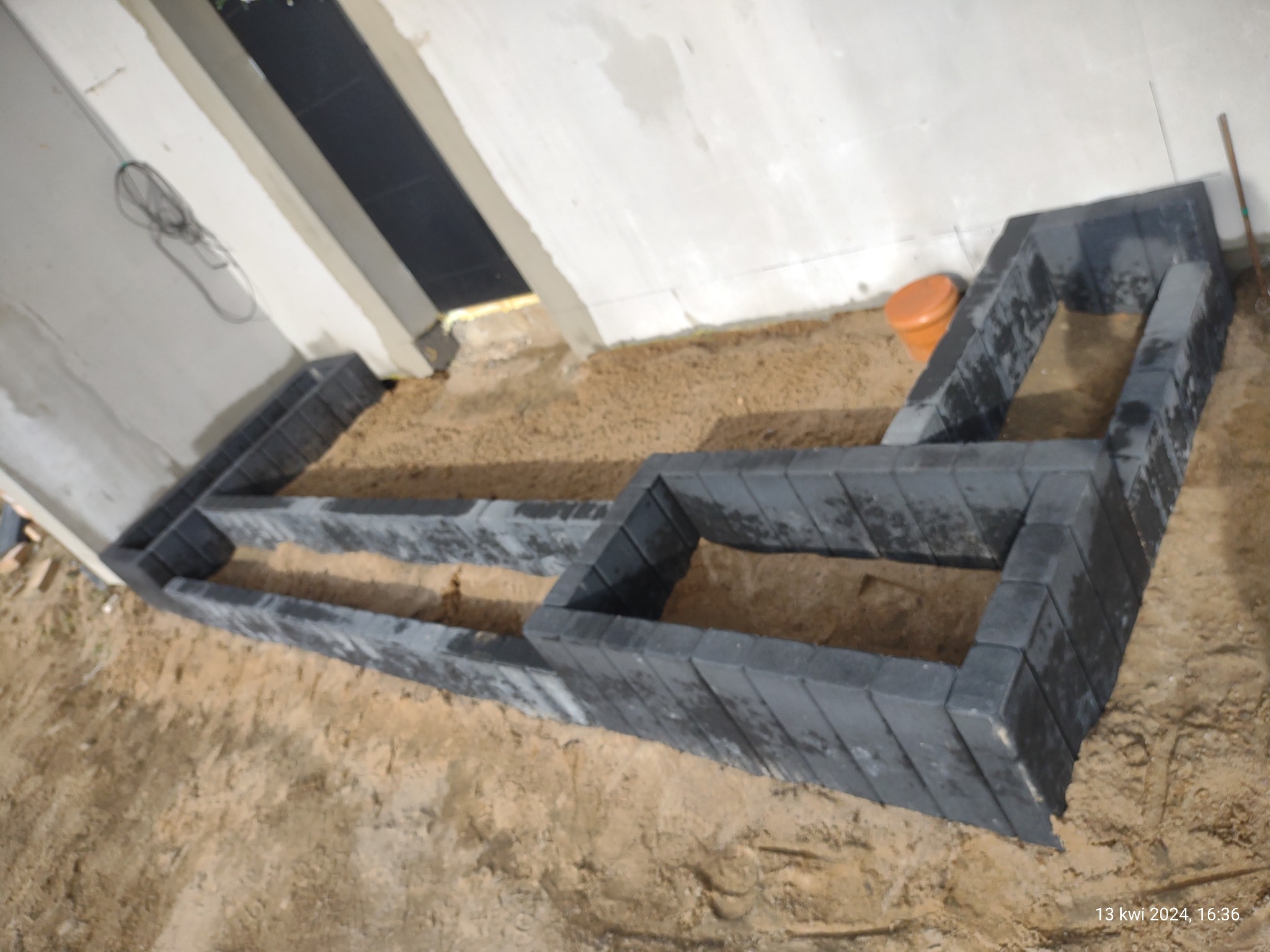 Przygotowanie podłoża pod taras z ciemnej kostki brukowej w trakcie budowy, widoczne warstwy piasku i obrzeża z kostki, surowe ściany budynku w tle.