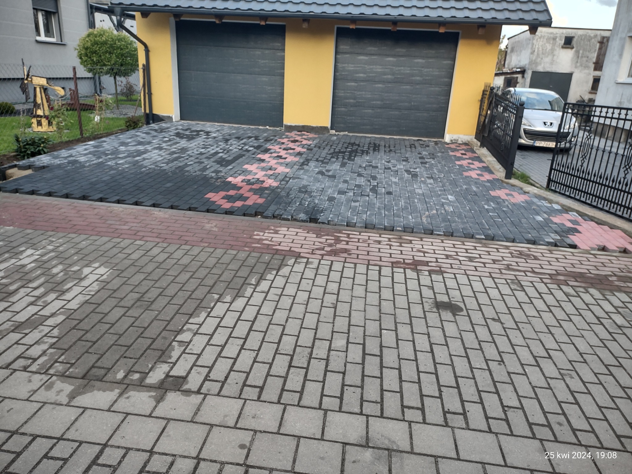 Podjazd do garażu wyłożony czarno-czerwono-szarą kostką brukową, na tle żółtej ściany garażu i częściowo widocznego samochodu osobowego za czarnym ogrodzeniem.