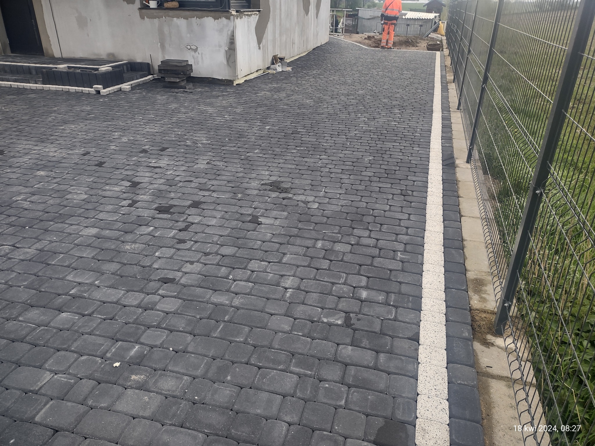 Ułożona, ciemnoszara kostka brukowa tworząca podjazd, z białym pasem oddzielającym od ogrodzenia z siatki. W tle widoczny budynek w trakcie wykończenia i pracownik w pomarańczowym ubraniu.