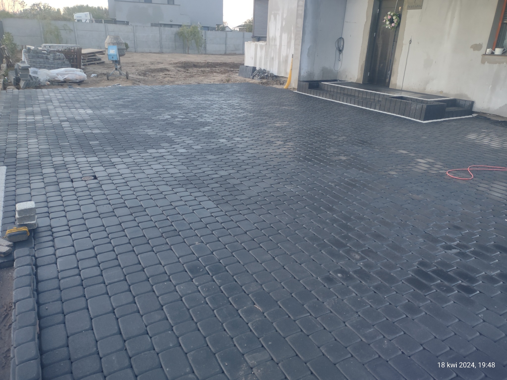Ułożona, mokra, grafitowa kostka brukowa przed nowoczesnym domem z ciemnymi drzwiami wejściowymi i schodami obłożonymi kostką, widoczne zapasy kostki i betoniarka.