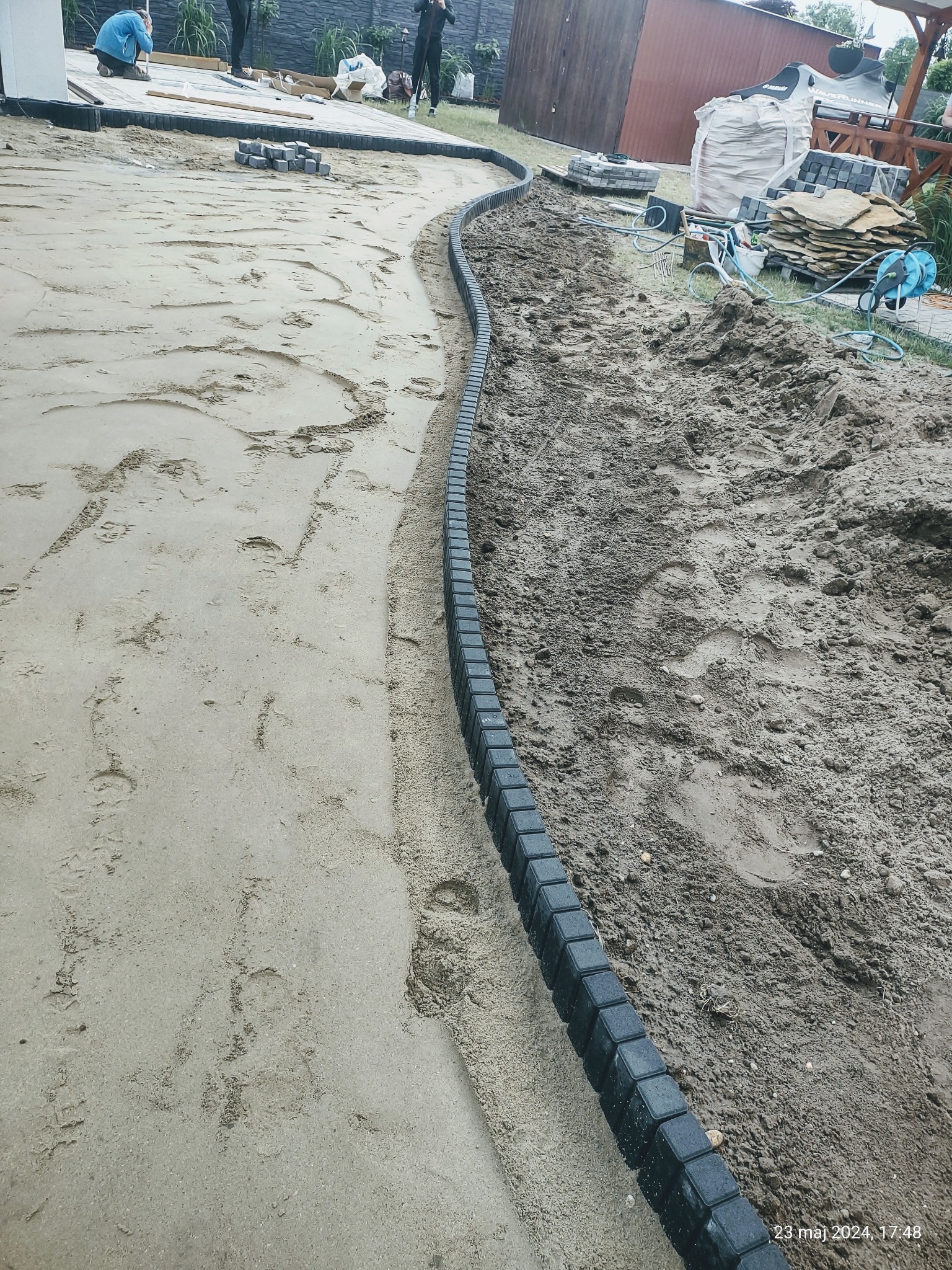 Przygotowanie podłoża pod ścieżkę z ciemnej kostki brzegowej; widoczne warstwy piasku i ziemi, oraz pracownicy w tle