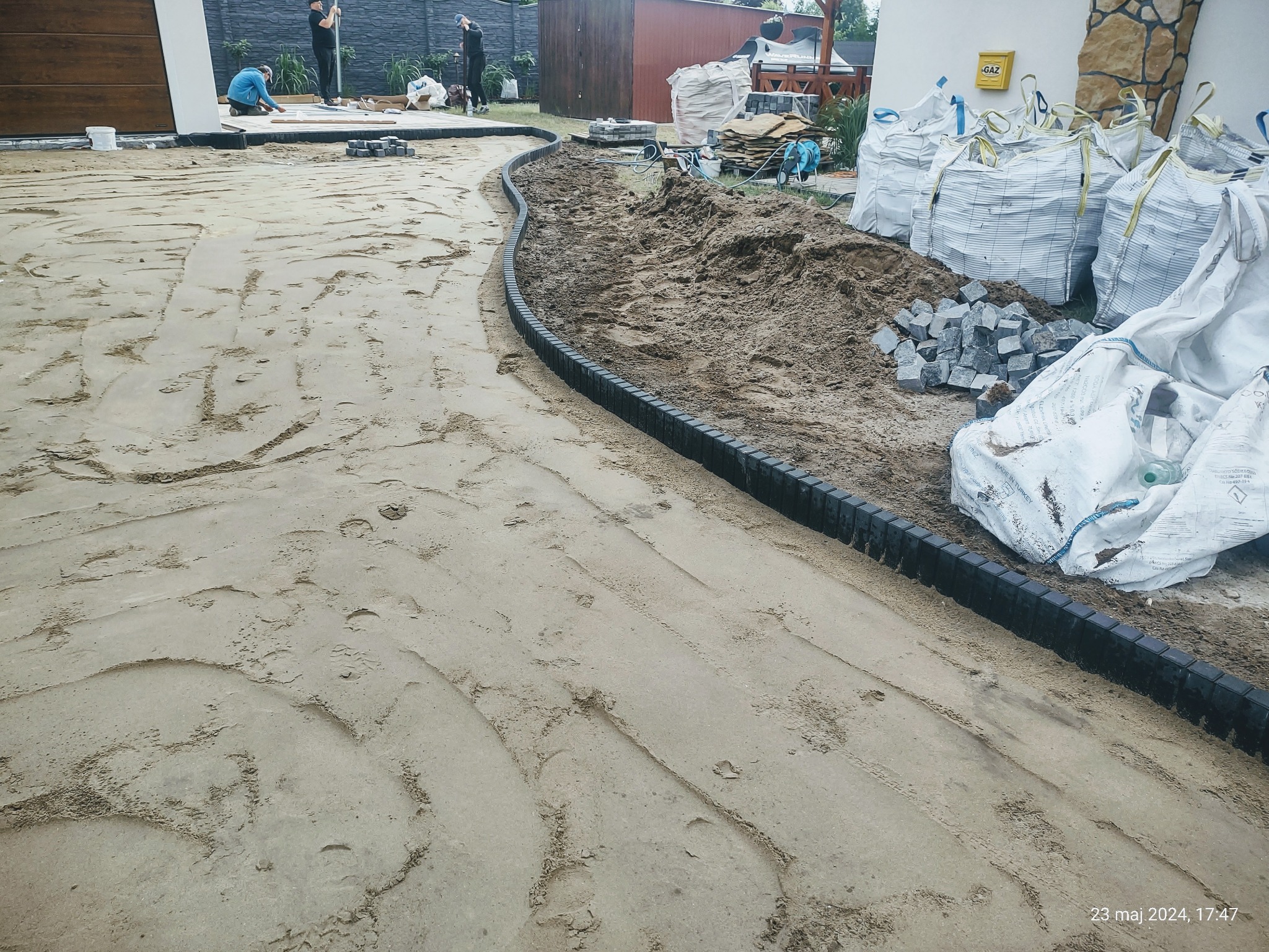 Przygotowanie podłoża pod kostkę brukową z ułożonym obrzeżem z tworzywa, widoczne worki z materiałem i ekipa budowlana w tle.